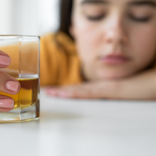 Can you die from a hangover? an image of a woman with her eyes closed, resting her head on her harm on a surface whilst holding an alcoholic beverage.